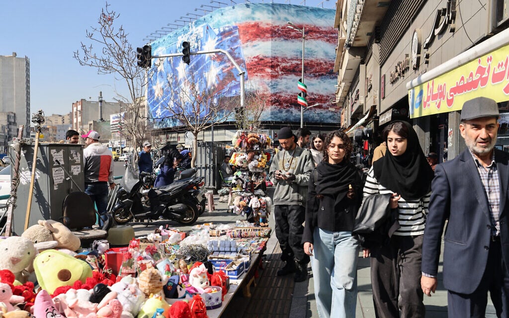 Iranians walk past a street vendor's stand with an anti-US billboard visible in the background on Enqelab Square in Tehran on February 17, 2026. (ATTA KENARE / AFP)