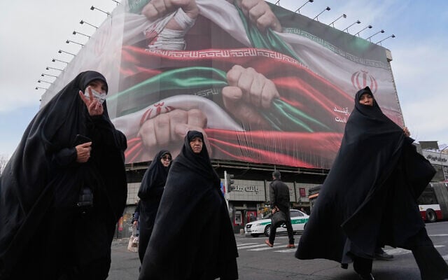 Women, one flashing a victory hand gesture, cross a street under a huge banner showing hands firmly holding Iranian national flags as a sign of patriotism, in Tehran, January 14, 2026. (AP Photo/Vahid Salemi) Women, one flashing a victory hand gesture, cross a street under a huge banner showing hands firmly holding Iranian national flags as a sign of patriotism, in Tehran, January 14, 2026. (AP Photo/Vahid Salemi)