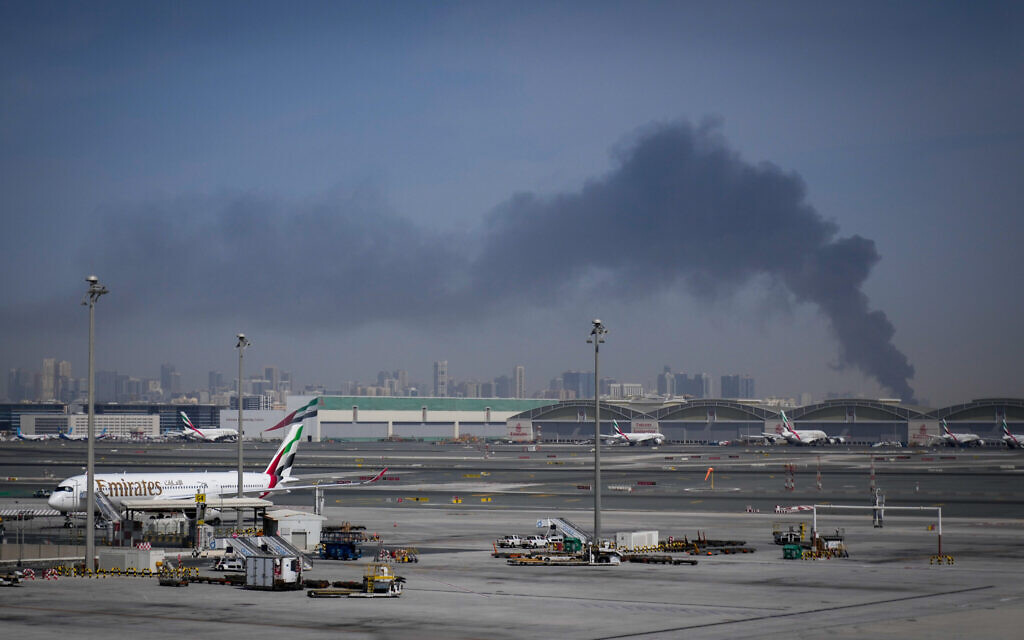 A plume of smoke caused by an Iranian strike is seen in the background as Emirates planes are parked at Dubai International Airport after its closure in Dubai, United Arab Emirates, Sunday, March 1, 2026. (AP/Altaf Qadri)
