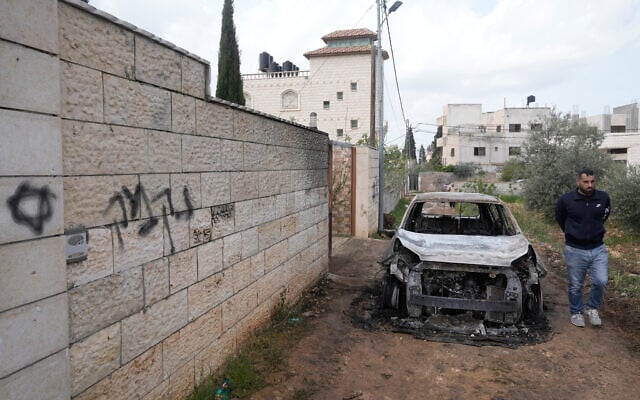A Palestinian man inspects a torched vehicle next to Hebrew graffiti on a wall that reads "revenge," following a settler rampage through nearby Nablus-area villages, in West Bank village of Deir al-Hatab, March 23, 2026. (AP/Majdi Mohammed) A Palestinian man inspects a torched vehicle next to Hebrew graffiti on a wall that reads "revenge," following a settler rampage through nearby Nablus-area villages, in West Bank village of Deir al-Hatab, March 23, 2026. (AP/Majdi Mohammed)