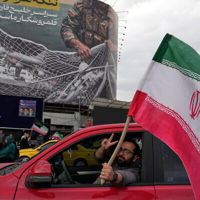 A man flashes a victory sign as he carries an Iranian flag in front of an anti-US billboard depicting the American aircrafts into the Iranian armed forces fishing net with signs that read in Farsi: "The Strait of Hormuz will remain closed, The entire Persian Gulf is our hunting ground," at the Eqelab-e-Eslami, or Islamic Revolution Square in downtown Tehran, Iran, April 13, 2026. (AP Photo/Vahid Salemi)