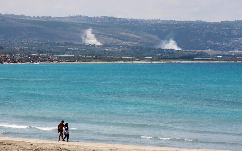 A man and woman walks along the beach a smoke rises following a couple of Israeli airstrikes targeting Hezbollah in the village of al-Qlailah, in southern Lebanon on May 25, 2026 (Kawnat HAJU / AFP)