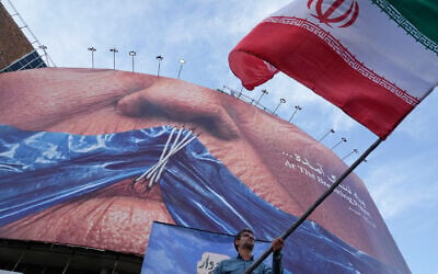 A man waves an Iranian flag for a pro-government campaign under a billboard with graphic showing Strait of Hormuz and sewn lips of US President Donald Trump in a square in downtown Tehran, Iran, on May 6, 2026. (AP Photo/Vahid Salemi)