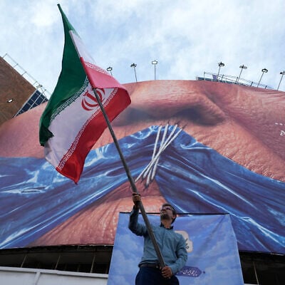 A man waves an Iranian flag for a pro-government campaign under a billboard with a graphic showing the Strait of Hormuz and sewn lips of US President Donald Trump in a square in downtown Tehran, May 6, 2026. (AP Photo/Vahid Salemi)