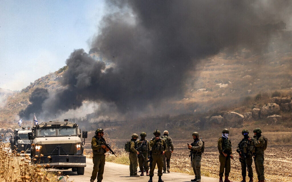 Israeli security forces stand guard along a road while behind smoke billows from an area reportedly set ablaze by Israeli settlers in the Palestinian village of Idna, west of Hebron in the West Bank, on June 5, 2026. (HAZEM BADER / AFP)