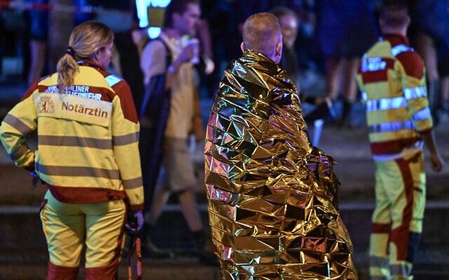 A man wrapped in a security blanket stands next to emercency personnel near the scene where one person was killed and around 15 injured after a car rammed into a crowd on the outskirts of the Christopher Street Day (CSD) parade in Berlin, on July 25, 2026. (RALF HIRSCHBERGER/AFP)