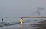 A woman stands at the water's edge along the Strait of Hormuz as a plume of smoke rises in the background following an explosion, off Bandar Abbas, Iran, July 13, 2026. (Razieh Poudat/ISNA via AP)