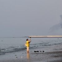 A woman stands at the water's edge along the Strait of Hormuz as a plume of smoke rises in the background following an explosion, off Bandar Abbas, Iran, July 13, 2026. (Razieh Poudat/ISNA via AP)