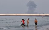 Three boys play in the shallow waters of the Strait of Hormuz, as a plume of smoke rises from an explosion in the background, off Bandar Abbas, Iran, July 13, 2026. (Razieh Poudat/ISNA via AP)