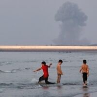 Three boys play in the shallow waters of the Strait of Hormuz, as a plume of smoke rises from an explosion in the background, off Bandar Abbas, Iran, July 13, 2026. (Razieh Poudat/ISNA via AP)