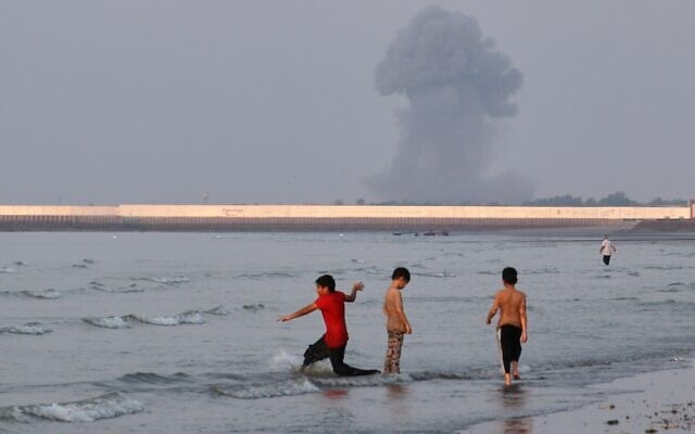 Three boys play in the shallow waters of the Strait of Hormuz, as a plume of smoke rises from an explosion in the background, off Bandar Abbas, Iran, July 13, 2026. (Razieh Poudat/ISNA via AP)