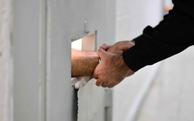 A guard grasps the hands of a cuffed prisoner through a door slot at the Givon Prison in Ramle on January 11, 2026. (Oren Cohen/Flash90)