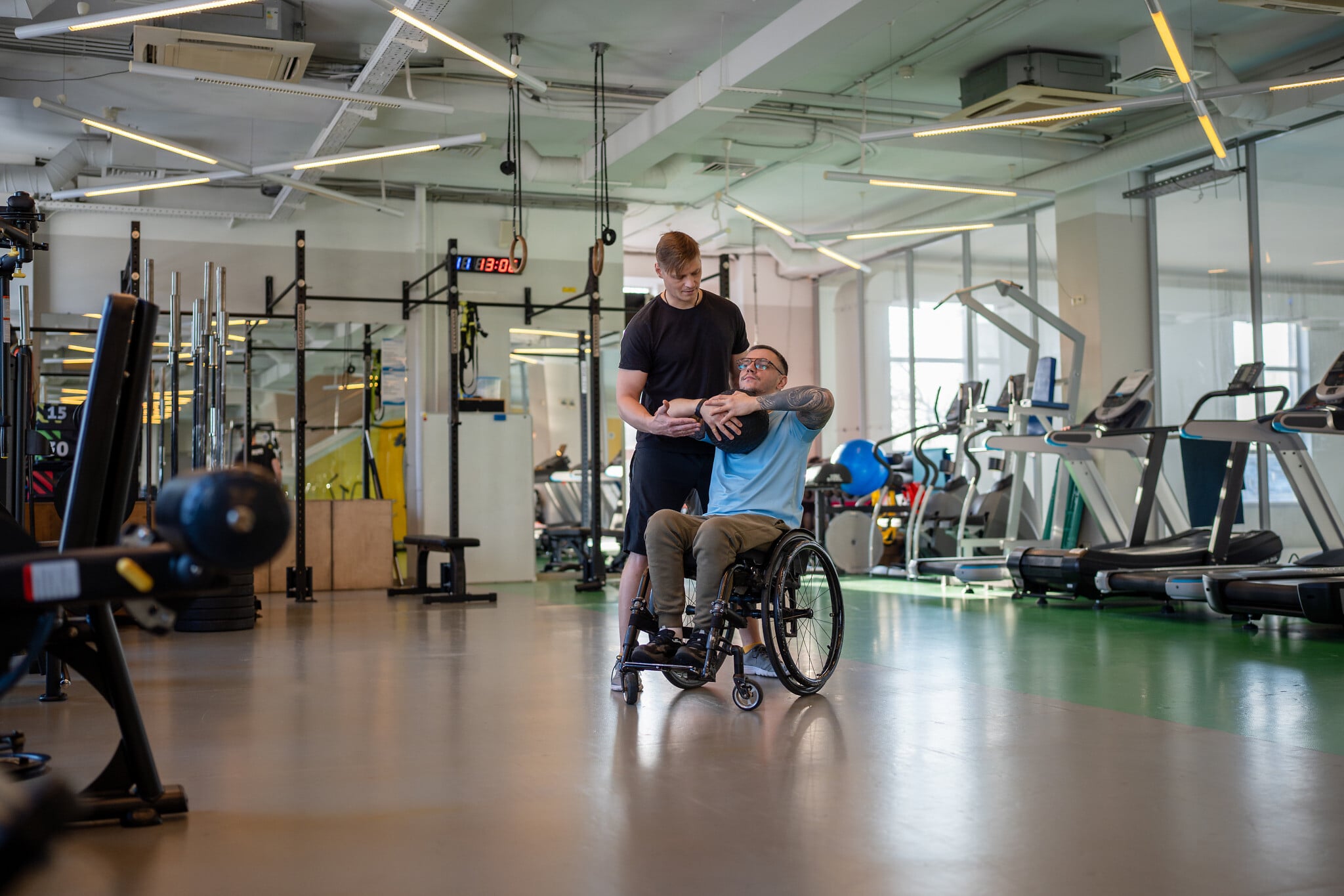 Personal sports trainer assists disabled sportsman in wheelchair with medicine ball workout to build upper body strength. Athlete undergoing rehabilitation in sports center with professional coach. Personal sports trainer assists disabled sportsman in wheelchair with medicine ball workout to build upper body strength. Athlete undergoing rehabilitation in sports center with professional coach.