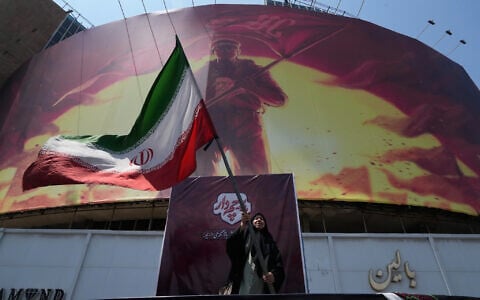A woman waves an Iranian flag next to a billboard showing a graphic of a man wearing a shirt showing the face of late Iranian supreme leader Ayatollah Ali Khamenei with words calling upon the avengers of Imam Hussein, in a square in downtown Tehran, Iran, August 1, 2026. (AP Photo/Vahid Salemi)