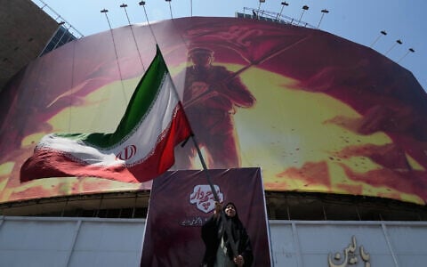 A woman waves an Iranian flag beneath a billboard showing a graphic calling for revenge on Imam Hussein, one of Shiite Islam’s most beloved saints, who was killed in a 7th century battle, and the late Iranian supreme leader Ayatollah Ali Khamenei, who was killed in a US and Israel strike on February 28, in a square in downtown Tehran, Iran, August 1, 2026. (AP Photo/Vahid Salemi)