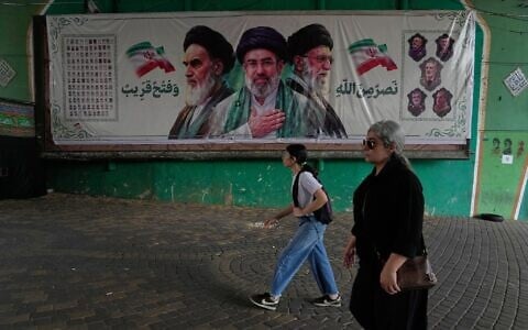Women walk past a banner showing portraits of the late Iranian revolutionary founder Ayatollah Khomeini, left, Supreme Leader Ayatollah Mojtaba Khamenei, center, and the late supreme leader Ayatollah Ali Khamenei, who was killed in the US and Israel strike on February 28, under a bridge in northern Tehran, Iran, August 2, 2026. (AP Photo/Vahid Salemi)