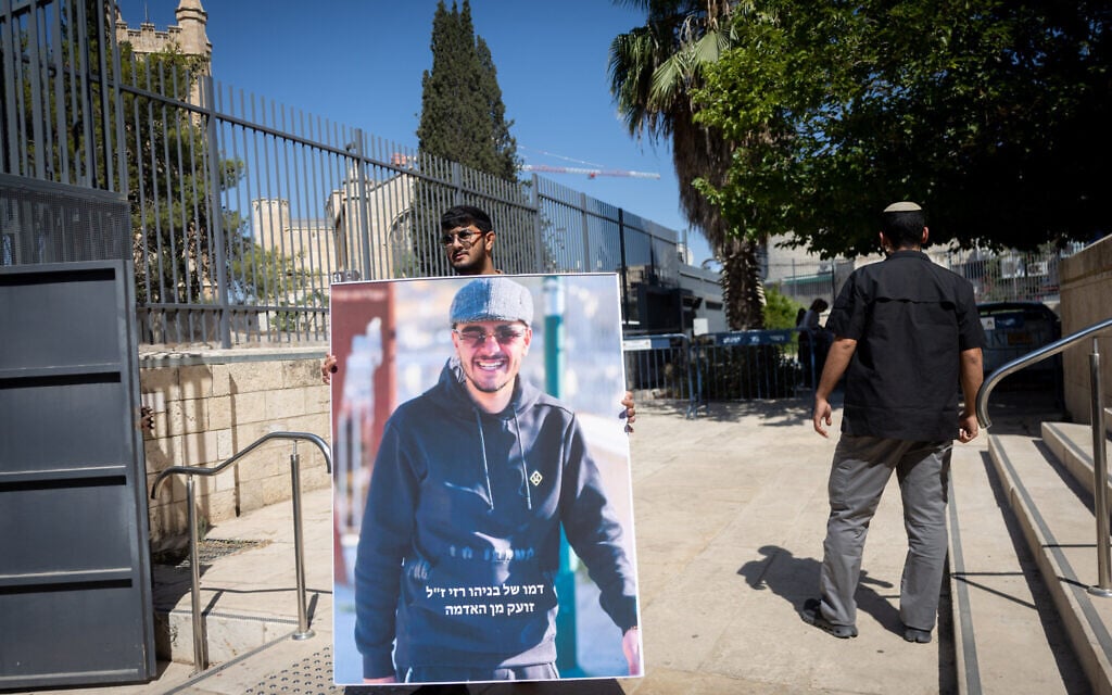 A young man holds an picture of Benayahu Razi who was murdered last month in Jerusalem, outside a court hearing on August 7, 2026. (Chaim Goldberg/FLASH90)