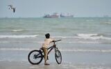 A gull flies overhead as a boy stands with his bicycle on a beach overlooking commercial vessels anchored in the Strait of Hormuz off Bandar Abbas, Iran, September 7, 2026. (AP Photo/Vahid Salemi)