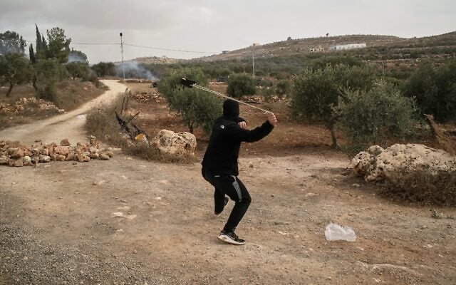 A Palestinian uses a sling to hurl a stone as tear gas drifts among olive trees during clashes following an alleged settler attack on Palestinians harvesting olives in the West Bank village of Abu Falah, October 16, 2026. (AP Photo/Leo Correa)